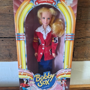 May include: A vintage-style doll in a red jacket with a white shirt, blue skirt, and blonde ponytail hair. The doll is holding a football and has a number 5 on her jacket. The box features the text "Bobby Sox" and "The Fun & Fabulous Fifties are Back!"
