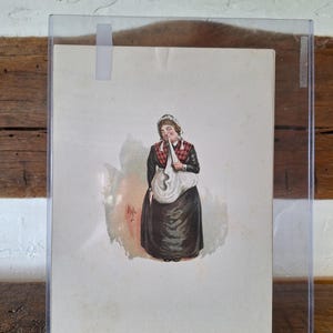 Può includere: Una stampa antica incorniciata con una caricatura colorata di una donna con un vestito nero e un grembiule bianco. La donna tiene un panno bianco davanti al viso. La stampa è in una custodia di plastica trasparente, montata su una superficie di legno.