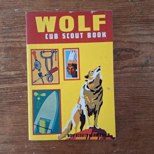 May include: Vintage "Wolf Cub Scout Book" with a yellow cover. The cover features the word "WOLF" in large yellow letters above "CUB SCOUT BOOK" in red. Illustrations include tools, a Native American profile, a boat, and a howling wolf.