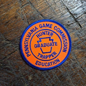 Może przedstawiać: Okrągła pomarańczowo-niebieska naszywka z napisem "Pennsylvania Game Hunter Commission Graduate Trapper Education". Naszywka ma niebieską obwódkę i leży na drewnianej powierzchni.