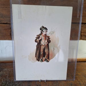 Può includere: Una stampa antica di un uomo con cappotto marrone, pantaloni a quadri e sciarpa rossa, che fuma la pipa. L'opera d'arte è in una custodia di plastica trasparente, esposta su una superficie di legno. Il testo "THE ARTFUL DODGER" è in basso.