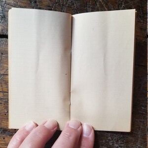 May include: A blank, cream-colored, small notebook with a sewn binding. The notebook is open and the pages are blank.