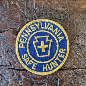 Könnte beinhalten: Runder Aufnäher mit den Worten "PENNSYLVANIA SAFE HUNTER" in goldener Schrift auf blauem Grund. In der Mitte befindet sich ein goldenes Schlüsselsymbol mit einem Kreuz. Goldener Rand.