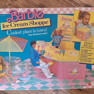 Può includere: Un set di gioco Barbie Ice Cream Shoppe rosa e giallo con oltre 50 pezzi. Il set include un carrello per gelati rosa, un bancone giallo, due sgabelli, due sedie, un tavolo, un ombrello e due bambole. La scatola presenta il testo "Barbie Ice Cream Shoppe" e "Coolest place in town!"