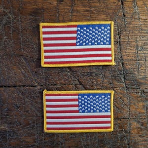 Può includere: Due toppe rettangolari raffiguranti la bandiera americana. Ogni toppa ha un cantone blu con stelle bianche, strisce rosse e bianche e un bordo giallo. Le toppe sono presentate su una superficie di legno rustico.