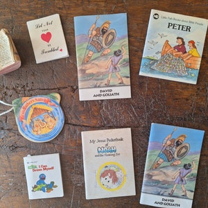 Puede incluir: Una colección de pequeños libros y adornos con temas religiosos. Los libros incluyen títulos como "David y Goliat" y "Pedro". Los adornos incluyen una escena de la natividad y un pequeño libro con el texto "Puedo vestirme solo".