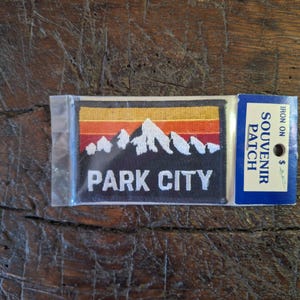 Peut inclure: Un écusson souvenir thermocollant rectangulaire avec un motif de chaîne de montagnes aux couleurs du coucher de soleil. Le patch porte l'inscription "PARK CITY" en lettres blanches. L'écusson est emballé dans un sac en plastique transparent.
