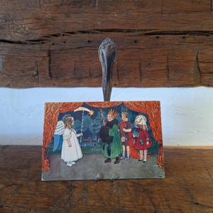 May include: A vintage rectangular postcard featuring a colorful illustration of an angel holding a star wand and three figures in medieval attire, held upright by a silver-toned metal spoon handle against a rustic wooden background.