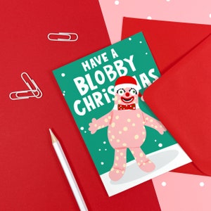 May include: A green Christmas card with a pink, polka-dotted character wearing a red hat and a red bow tie. The card says "Have a Blobby Christmas".