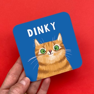 May include: Square coaster with a blue background and the word "DINKY" in white. The coaster features a cartoon illustration of an orange tabby cat with green eyes.