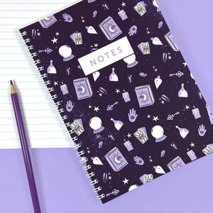 Peut inclure: Carnet à spirales violet avec une couverture violet foncé ornée d'un motif répété d'étoiles, de cristaux, de clés, de fioles de potion, de mains et d'un croissant de lune. Le texte "NOTES" est imprimé en blanc sur un rectangle blanc au centre de la couverture.