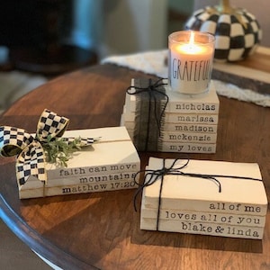 May include: Three stacks of decorative wooden books with text and black twine. One stack has a black and white checkered bow and greenery. A lit candle with the word "GRATEFUL" sits atop another stack. The books have names and phrases.