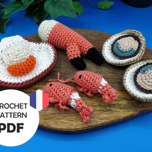 Puede incluir: Patrón de crochet PDF para un conjunto de mariscos amigurumi que incluye camarones y ostras. El patrón está disponible en francés. Los amigurumi se muestran en una tabla de cortar de madera.