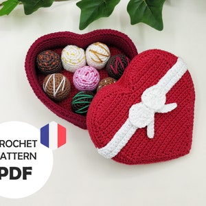 May include: A crocheted heart-shaped box with a white bow and lid open to reveal a collection of crocheted chocolate candies inside.