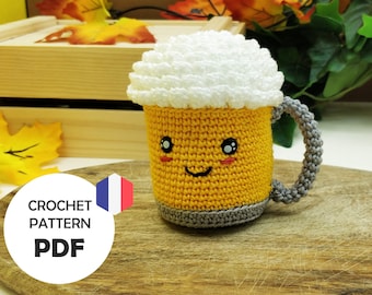 May include: A crocheted beer mug with a white foam top and a smiling face. The mug is yellow with a gray handle and is sitting on a wooden surface.