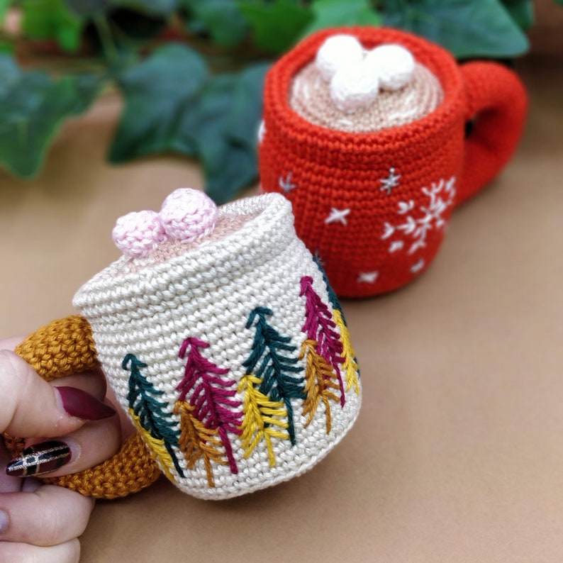 以下が含まれることがあります： 刺繍が施された2つの編み物製のマグカップ。1つのマグカップは白の雪片がついた赤色で、もう1つは森のシーンがついたクリーム色です。両方のマグカップは茶色の取っ手があり、白いマシュマロでいっぱいです。
