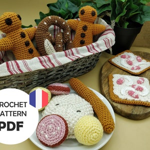 以下が含まれることがあります： 編みぐるみのおもちゃの食べ物セットのクロシェパターンPDF。ジンジャーブレッドマン、プレッツェル、ソーセージ、パン、チーズボール、ピンクのジャムを塗ったパン2枚が含まれています。おもちゃは籐のバスケットと白い皿に入っています。