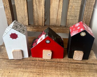 May include: Three miniature wooden houses decorated for Christmas. The houses are painted white, red, and black and have different patterned paper on the roof. Each house has a small wooden door and a snowflake sticker on the front.
