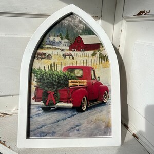 May include: A white framed picture of a red pickup truck carrying a Christmas tree. The truck is driving through a snowy landscape with a red barn in the background. The truck has a sign that says "Apple Valley Orchards Nursery & Tree Farm".