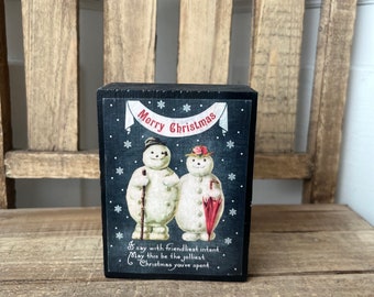 May include: A black wooden block with a vintage Christmas card image of two snowmen. The card has a white banner with the text "Merry Christmas" and a message that reads "I say with friendliest intent. May this be the jolliest Christmas you've spent."