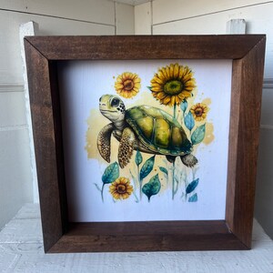 May include: A framed watercolor painting of a green sea turtle swimming through a field of yellow sunflowers. The turtle is facing the right side of the image and has a light brown shell with dark brown markings. The sunflowers are in various stages of bloom, with some fully open and others still in bud.
