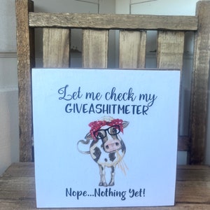 May include: A white wooden sign with black text that reads "Let me check my giveashitmeter Nope...Nothing Yet!"  A cartoon cow with black and white spots is wearing red and white polka dot glasses and a red bandana.