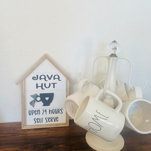 May include: A white wooden sign with the text "Java Hut" and a black silhouette of a coffee cup with a heart. The sign also says "Open 24 Hours Self Serve". A white ceramic mug with the word "Home" in black lettering is on a white metal mug rack.