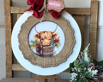 May include: A round, white and wood-framed wall hanging with a Highland cow image. The cow is in a basket with flowers. A red bow and a tag that says "Winter Magic" are attached to the top.