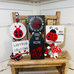 May include: A collection of ladybug-themed wooden signs with sayings like "Sweet Summer Shine Your Light", "Every Bug Welcome", "Ladybug Crossing", and "Hello Summer". The signs are displayed on a small wooden bench with red and white beads and a red and white mushroom.