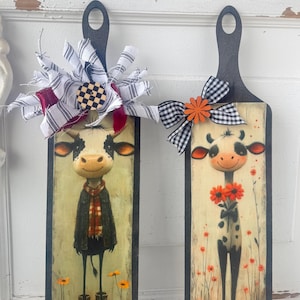May include: Two decorative wooden cutting boards with black handles. Each board features a whimsical cow illustration. One cow wears a scarf and boots, the other holds flowers. Both boards have decorative bows.