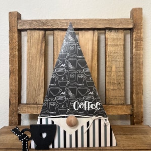 May include: Black and white coffee cup gnome with a black and white striped hat and a wooden nose. The gnome is sitting on a wooden surface. The gnome's hat has the word "Coffee" written on it.