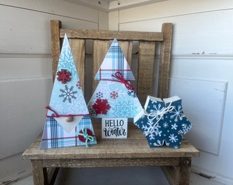May include: Three wooden Christmas tree ornaments decorated with blue and white snowflake patterns, red plaid, and red snowflakes. The center tree has a sign that reads "Hello Winter".