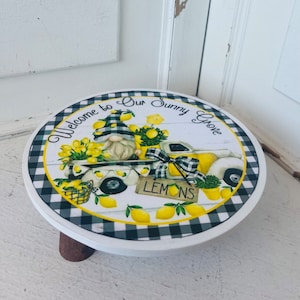 May include: A white round wooden stool with a black and white checkered border. The top of the stool features a gnome driving a yellow truck with lemons and a sign that reads "Lemons". The text "Welcome to Our Sunny Grove" is written around the border.