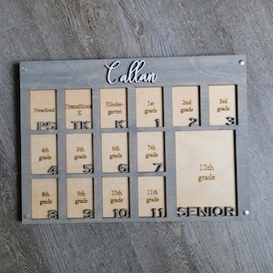 May include: A wooden photo frame with a gray background and a white inscription that reads "Callan". The frame has 12 slots for photos, each labeled with a grade level from preschool to 12th grade. The frame is designed to hold photos of a child's progress through school.