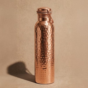 May include: A cylindrical copper water bottle featuring a hammered texture across its body. The bottle has a smooth, flat base and a matching copper screw-top lid with a thin, integrated metal loop handle attached to the side of the cap.
