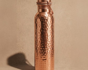 May include: A cylindrical copper water bottle featuring a hammered texture across its body. The bottle has a smooth, flat base and a matching copper screw-top lid with a thin, integrated metal loop handle attached to the side of the cap.