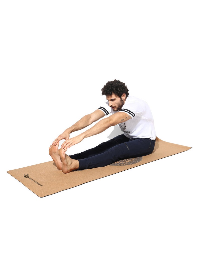 May include: A person stretches on a brown cork yoga mat with the text "Sakti Warrior" printed on it.