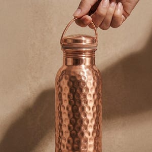 May include: A cylindrical copper water bottle featuring a distinct hammered texture across its surface. The bottle includes a matching metallic screw-top lid equipped with a thin, arched carrying handle attached to the neck for easy transport.
