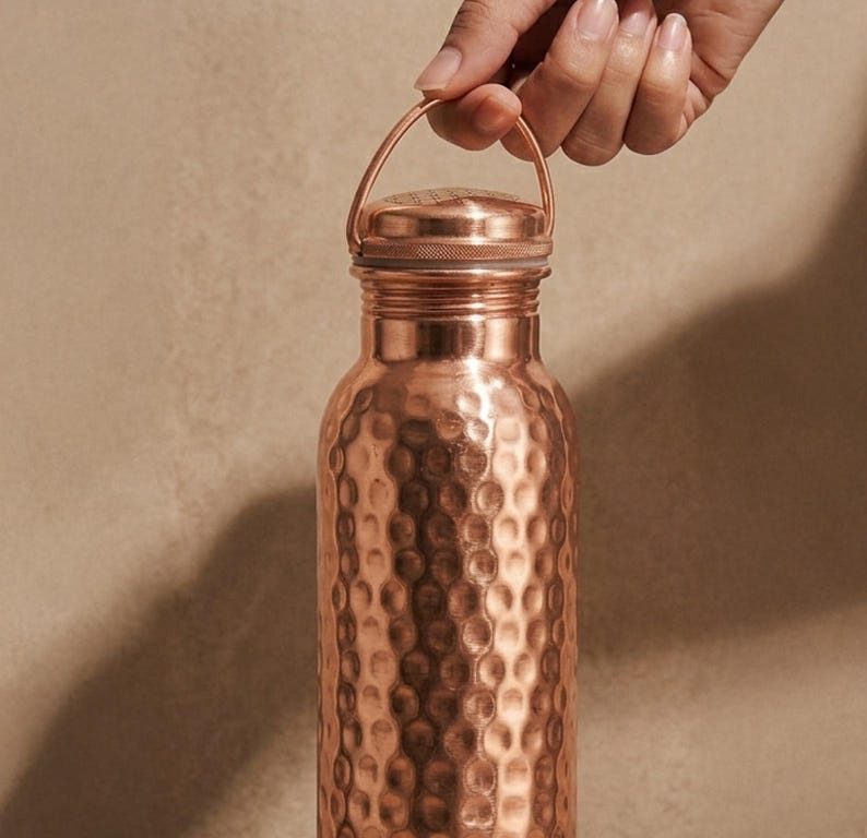 May include: A cylindrical copper water bottle featuring a distinct hammered texture across its surface. The bottle includes a matching metallic screw-top lid equipped with a thin, arched carrying handle attached to the neck for easy transport.