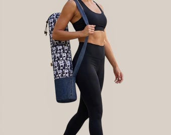 Puede incluir: Una bolsa cilíndrica para esterilla de yoga con un estampado de elefantes en blanco y negro en la parte superior y una base de tela azul oscuro tipo mezclilla, equipada con una correa larga y ajustable para llevarla al hombro.