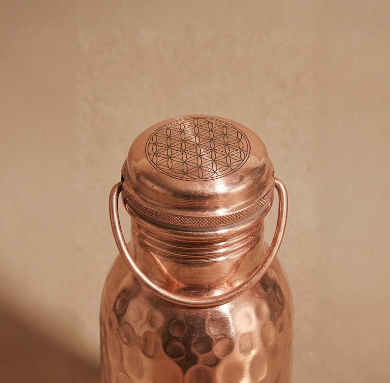 May include: A copper water bottle with a hammered texture and a round screw-top lid. The top of the lid features a laser-engraved geometric flower of life pattern. A thin, rounded copper handle is attached to the neck of the bottle.