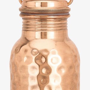 May include: A copper water bottle featuring a hammered texture on the main body, a smooth neck with horizontal ridges, and a circular screw-top lid with a thin metal handle. The lid displays a subtle engraved pattern on its top surface.
