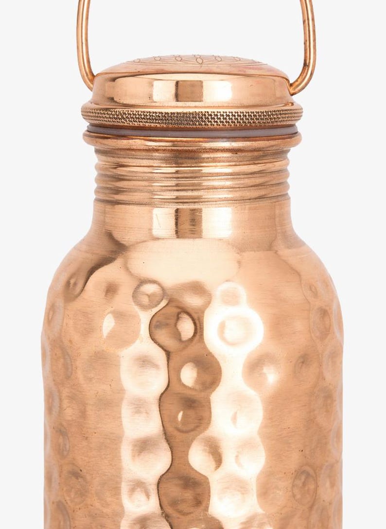 May include: A copper water bottle featuring a hammered texture on the main body, a smooth neck with horizontal ridges, and a circular screw-top lid with a thin metal handle. The lid displays a subtle engraved pattern on its top surface.