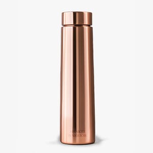May include: A copper-coloured water bottle with a screw-on cap. The bottle has a sleek, cylindrical design with a slightly wider base. The words "SHAKTI WARRIOR" are printed vertically near the bottom of the bottle.