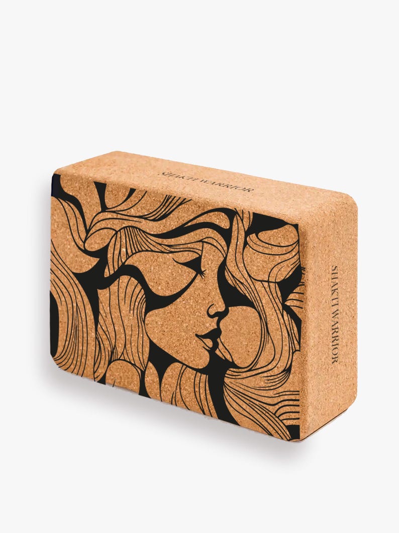 K&ouml;nnte beinhalten: Ein Kork-Yogablock mit einer schwarz-wei&szlig;en Illustration eines Frauenkopfes mit wallendem Haar. Der Text "Shakti Warrior" ist auf der Seite des Blocks aufgedruckt.