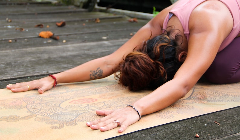 Puede incluir: Una persona practicando yoga en una esterilla con estampado. La persona lleva un top rosa y leggings morados. La esterilla es marr&oacute;n con un dise&ntilde;o de mandala colorido.