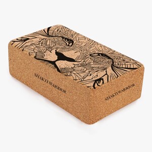 K&ouml;nnte beinhalten: Ein Kork-Yogablock mit einer Schwarz-Wei&szlig;-Zeichnung eines L&ouml;wenkopfes. Der Block ist rechteckig und hat auf zwei Seiten den Text "SHAKTI WARRIOR" aufgedruckt.
