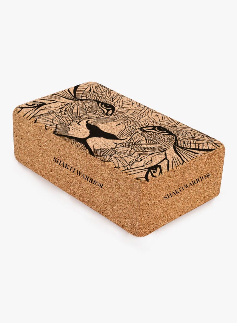 K&ouml;nnte beinhalten: Ein Kork-Yogablock mit einer Schwarz-Wei&szlig;-Zeichnung eines L&ouml;wenkopfes. Der Block ist rechteckig und hat auf zwei Seiten den Text "SHAKTI WARRIOR" aufgedruckt.