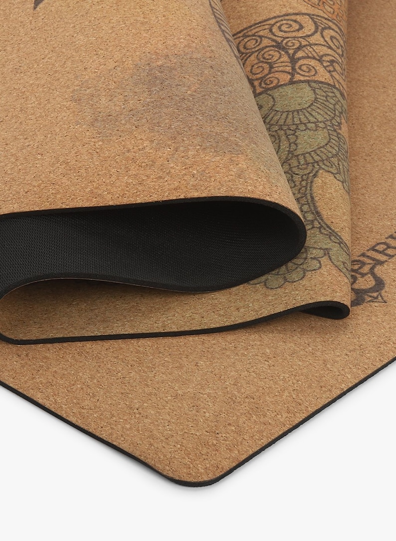 Puede incluir: Una esterilla de yoga de corcho marr&oacute;n con un borde negro y un dise&ntilde;o de mandala verde y marr&oacute;n. La esterilla est&aacute; enrollada y el dise&ntilde;o es visible en el exterior del rollo.