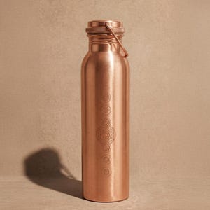 May include: A cylindrical copper water bottle with a metallic finish and a screw-top lid featuring a thin metal loop handle. The front of the bottle is engraved with a vertical series of seven circular chakra symbols arranged in a linear pattern.